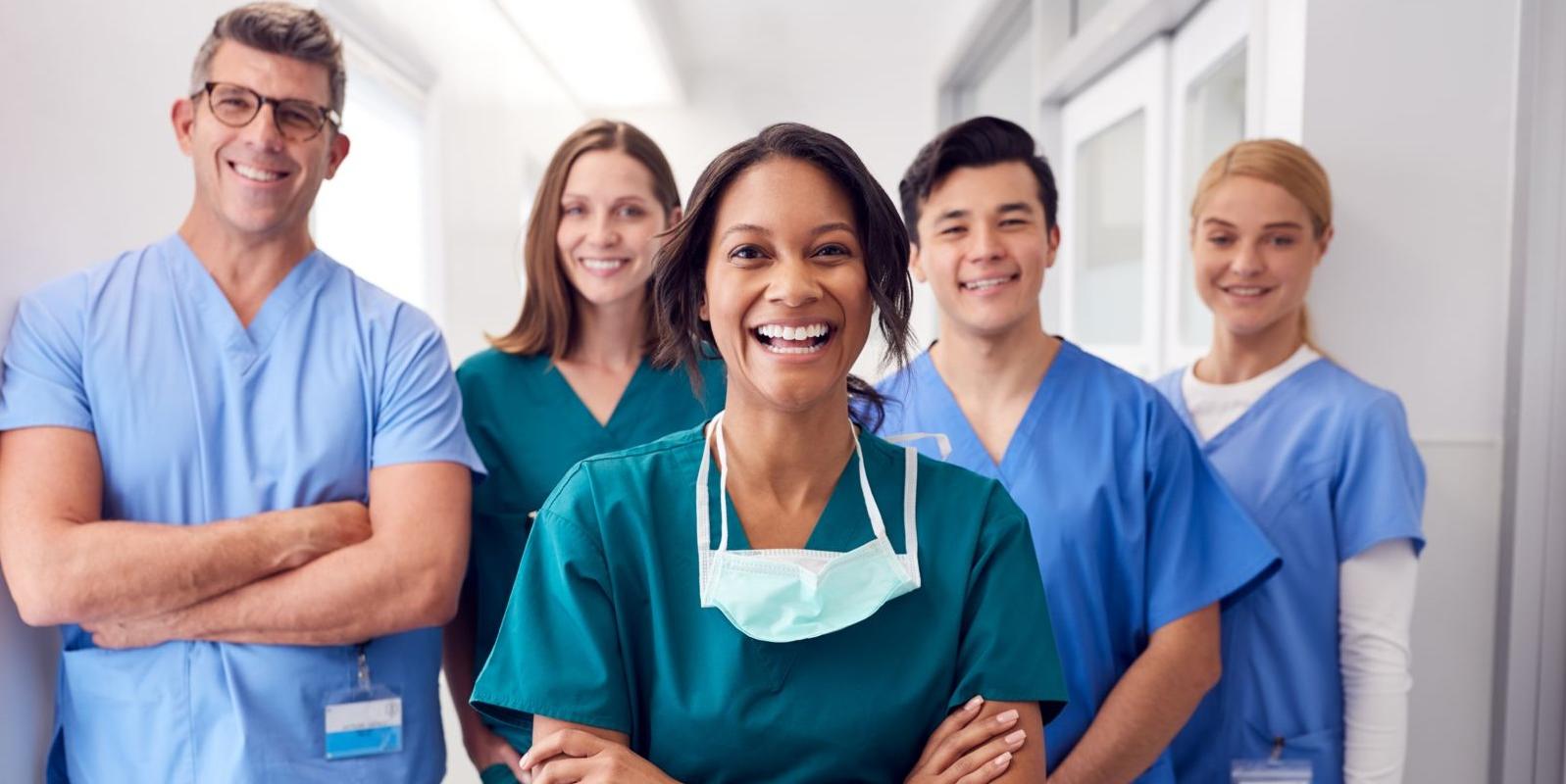 Five health care workers stand in a hallway wearing scrubs in various shades of blue. An African American female stands center with a Caucasian male and female standing directly behind her. Another Caucasian female stands to the far left behind the Caucasian male and another tall Caucasian male stands to the far right.